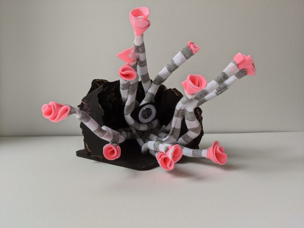 A small sculpture made from found objects looking kind of like an octopus but with one big eye in the centre of their body. They are emerging from a sculpture of a cave.